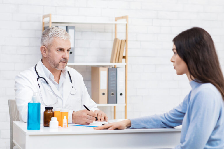 Problemy zdrowotne, choroby, b&oacute;l i recepty. Poważny starszy mężczyzna w białym fartuchu słucha pacjentki i robi notatki przy stole z lekami w słoikach i środkiem dezynfekującym we wnętrzu gabinetu.